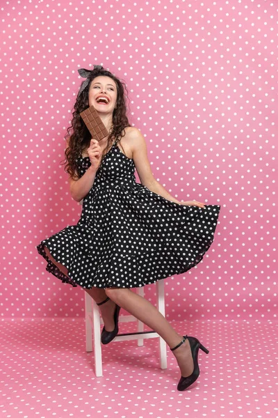 Nettes Mädchen mit welligem dunklem Haar posiert auf dem Stuhl mit einer Tafel Milchschokolade in den Händen, lacht verzweifelt und schaut zur Seite. — Stockfoto