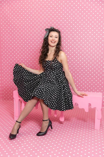 Chica caucásica rizado está sentado en el banco de color rosa, posando en la cámara, sonriendo, levantando un lado de su vestido . —  Fotos de Stock