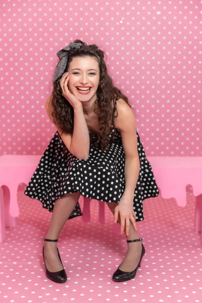 Curly menina caucasiana está sentado no banco rosa, posando para a câmera, sorrindo, inclinando-se para a frente . — Fotografia de Stock