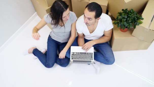 Paar zitten op de vloer van hun nieuwe flat en het maken van online winkelen op laptop, het kiezen van meubels en decorelementen. — Stockvideo