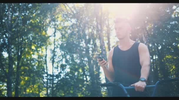 Mann sitzt auf BMX-Rad in Skatepark und benutzt Handy Die Sonne verschwimmt. Kopierraum. 4K. — Stockvideo