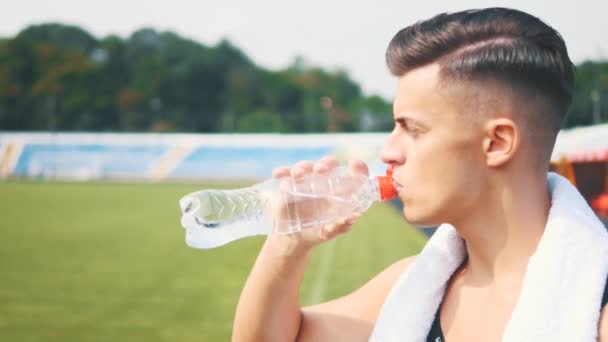 Il giovane sportivo sta finendo il suo allenamento allo stadio e bevendo acqua. Vista laterale. Chiudete. Ricevuto. 4K . — Video Stock