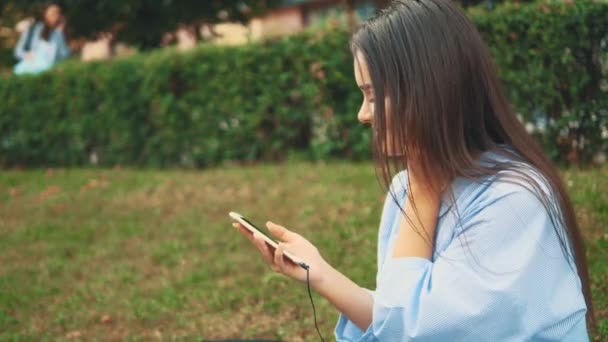 Menina bonita e adorável ouve música usando fones de ouvido no parque. Fecha. Vista lateral. Espaço para cópia. 4K . — Vídeo de Stock