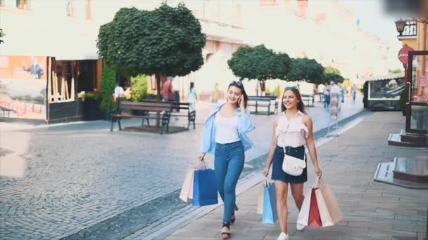 Dos mujeres jóvenes atractivas están caminando y hablando en una calle de verano con bolsas de compras. Uno de ellos está al teléfono. Lento. En cámara lenta. De cerca. Copiar espacio. 4K — Vídeos de Stock