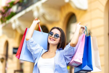 Young caucasian woman in sunglasses raising multi-coloured bags with purchases walking on the streets of shopping centre.