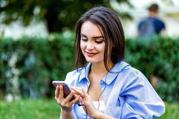 Chica encantadora sentada escuchando música en el parque, mirando a la cámara sonriente, pacífica e inspirada . — Foto de Stock