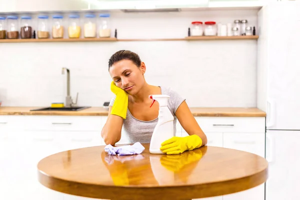 Woman got tired after hard work of cleaning, leaning on hand in protective glove and holding a bottle of spray over kitchen background.