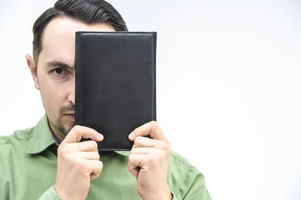 En ung man gömmer halva ansiktet bakom en svart bok, tittar beslutsamt på kameran, som en analytiker. — Stockfoto