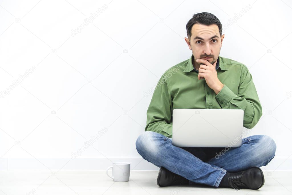 Casual businessman working, sitting in lotos position on the floor, typing on keyboard, looking at computer screen with concentrated face expression.