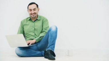 Yakışıklı genç adam dizinde dizüstü bilgisayarla yerde oturuyor ve yanında beyaz bir bardak, dizüstü bilgisayarında komik bir şeye bakıyor, konuşuyor ve kameraya gülümsüyor..