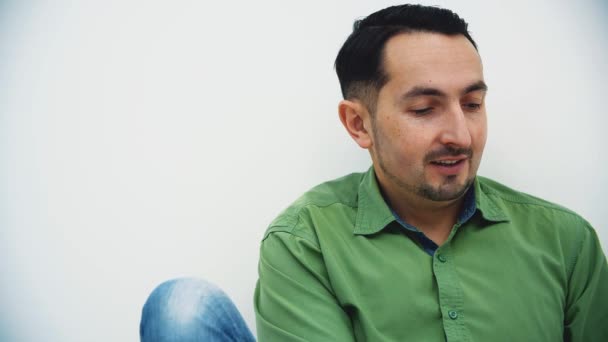 Joven guapo con camisa verde sentado en el suelo mirando hacia un lado con expresión de cara misteriosa . — Vídeos de Stock