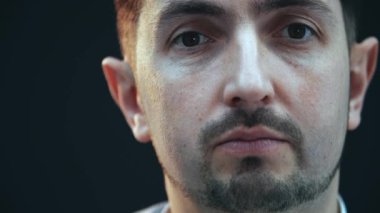 Ciddi bir genç adam, gözleri kapalı, kameraya bakıyor, konuşuyor..