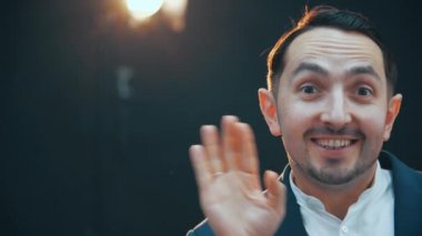 Yakışıklı, oyuncu, şık takım elbiseli genç adam ayakta duruyor, poz veriyor, gülümsüyor ve ellerini aptalca sallıyor, aptal yüz ifadeleri yapıyor, dudaklarını büküyor, kaşlarını çatıyor..