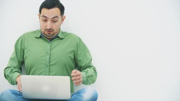 Odizolowany młody człowiek ze zdumioną głupią twarzą siedzący na podłodze w pozycji lotosu z laptopem na kolanach, oglądający coś szokującego, poruszający się szalonymi rękami. — Wideo stockowe