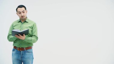 Adam ayakta duruyor, okuyor, defterinin sayfalarını karıştırıyor, sonra kameraya bakıyor, gülümsüyor..