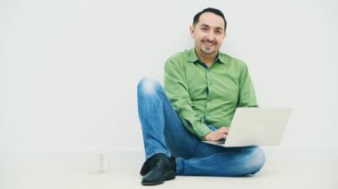 Genç adam dizinde dizüstü bilgisayarla yerde oturuyor ve yanında beyaz bir bardak, dizüstü bilgisayarında komik bir şey izliyor, dans ediyor, kameraya gülümsüyor..