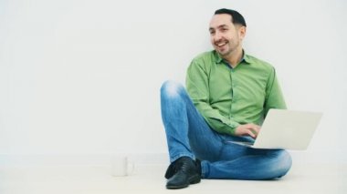 Yakışıklı genç bir adam dizinde dizüstü bilgisayarla yerde oturuyor ve yanında beyaz bir bardak, dizüstü bilgisayarında son derece komik bir şeye bakıyor, kameraya gülüyor..