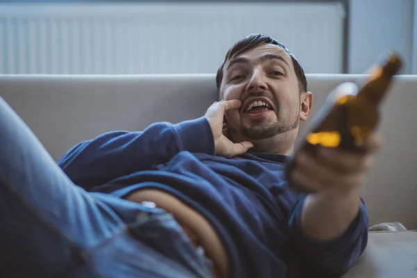 Mężczyzna leżący na kanapie proponuje butelkę piwa do picia.. — Zdjęcie stockowe
