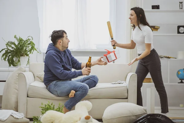 Um marido está bebendo cerveja enquanto sua esposa bate nele com um morcego . — Fotografia de Stock