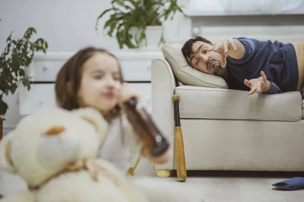 Little daughter trying to sip a beer while her dad is laying on a couch. — Stock Photo, Image