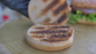 Profesyonel aşçıya yakın çekim, fast food restoranında lezzetli bir burger pişirir. Eldivenli kadın aşçı hamburgerler için üç gül ekmeğine ketçap koyar.