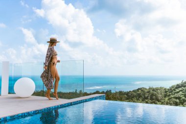 Mayo giymiş güzel bir kız, rüzgarda gelişen bir şapka ve bornoz, havuzu ve ormanın ve denizin güzel manzarası olan bir villada duruyor. Tayland, Samui