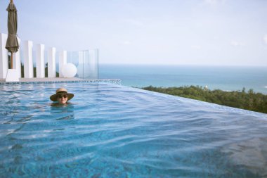 Mayo giymiş güzel bir kız, rüzgarda gelişen bir şapka ve bornoz, havuzu ve ormanın ve denizin güzel manzarası olan bir villada duruyor. Tayland, Samui