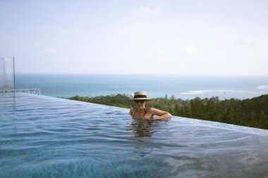 Mayo giymiş güzel bir kız, rüzgarda gelişen bir şapka ve bornoz, havuzu ve ormanın ve denizin güzel manzarası olan bir villada duruyor. Tayland, Samui