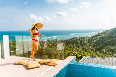 Kırmızı mayo giymiş güzel bir kız havuz kenarındaki lüks bir villada şarap ve güneşten korunan bir şapkayla dinleniyor. Palmiye ağaçları ve deniz manzarası. Tayland, Koh Samui