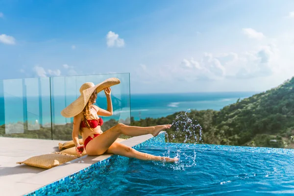 一个穿着红色游泳衣的漂亮姑娘正在游泳池边的一座豪华别墅里放松 她喝着酒 戴着一顶帽子遮遮掩掩着阳光 棕榈树和大海的景色 泰国Koh Samui — 图库照片