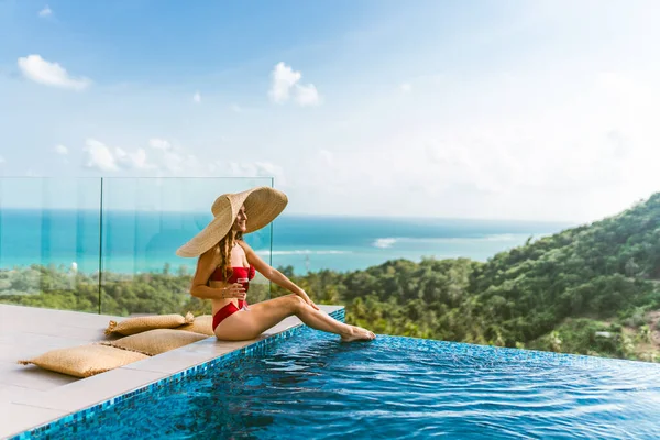 一个穿着红色游泳衣的漂亮姑娘正在游泳池边的一座豪华别墅里放松 她喝着酒 戴着一顶帽子遮遮掩掩着阳光 棕榈树和大海的景色 泰国Koh Samui — 图库照片
