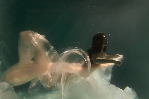 漂亮的姑娘留着长发在水里游泳 蓝色或金色背景如黄金 童话或魔法的气氛 用闪亮的布在水里潜水 图库图片