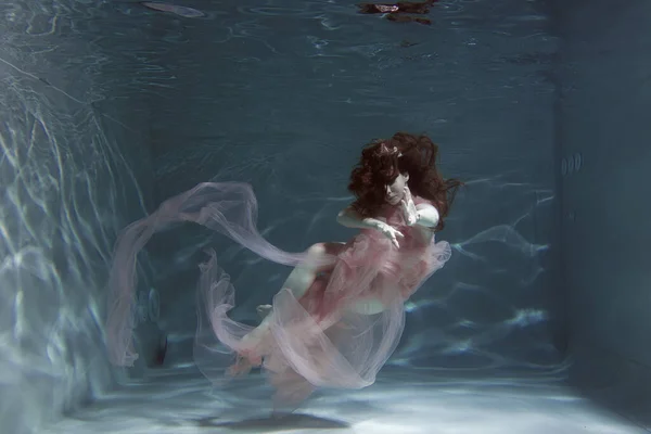 A girl with long dark hair swims underwater in a pink dress and with a crown on her head, like an underwater queen. Fairy tale suitable for advertising