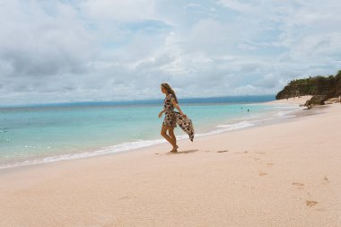 Okyanus ve denizin kıyısında mavi dalgalarla poz veren güzel ve genç bir kız. Uzun elbise mankeni ve moda pozu giymiş bir kadın. Bali adasında rahatla ve rahatla.