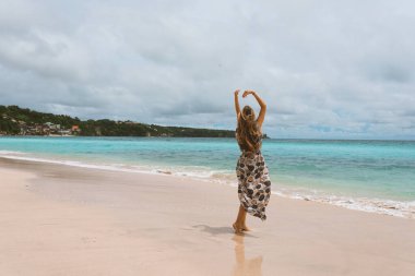 Okyanus ve denizin kıyısında mavi dalgalarla poz veren güzel ve genç bir kız. Uzun elbise mankeni ve moda pozu giymiş bir kadın. Bali adasında rahatla ve rahatla.