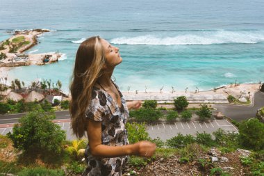 Okyanus ve denizin kıyısında mavi dalgalarla poz veren güzel ve genç bir kız. Uzun elbise mankeni ve moda pozu giymiş bir kadın. Bali adasında rahatla ve rahatla.