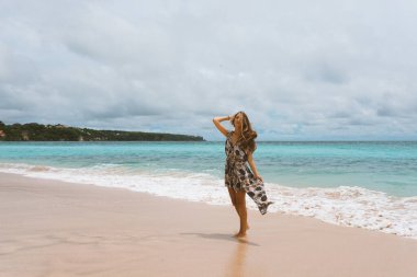 Okyanus ve denizin kıyısında mavi dalgalarla poz veren güzel ve genç bir kız. Uzun elbise mankeni ve moda pozu giymiş bir kadın. Bali adasında rahatla ve rahatla.