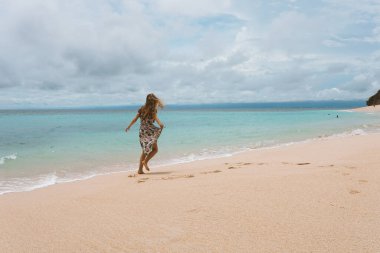 Okyanus ve denizin kıyısında mavi dalgalarla poz veren güzel ve genç bir kız. Uzun elbise mankeni ve moda pozu giymiş bir kadın. Bali adasında rahatla ve rahatla.