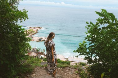 Okyanus ve denizin kıyısında mavi dalgalarla poz veren güzel ve genç bir kız. Uzun elbise mankeni ve moda pozu giymiş bir kadın. Bali adasında rahatla ve rahatla.