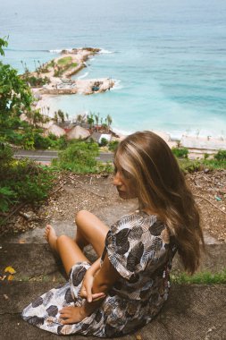 Okyanus ve denizin kıyısında mavi dalgalarla poz veren güzel ve genç bir kız. Uzun elbise mankeni ve moda pozu giymiş bir kadın. Bali adasında rahatla ve rahatla.