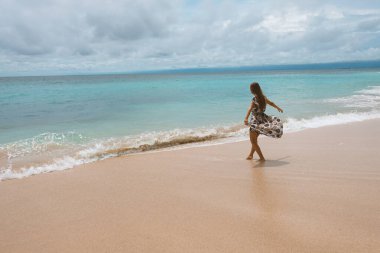 Okyanus ve denizin kıyısında mavi dalgalarla poz veren güzel ve genç bir kız. Uzun elbise mankeni ve moda pozu giymiş bir kadın. Bali adasında rahatla ve rahatla.