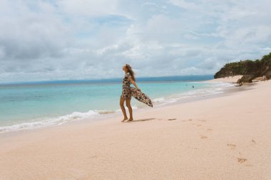 Okyanus ve denizin kıyısında mavi dalgalarla poz veren güzel ve genç bir kız. Uzun elbise mankeni ve moda pozu giymiş bir kadın. Bali adasında rahatla ve rahatla.
