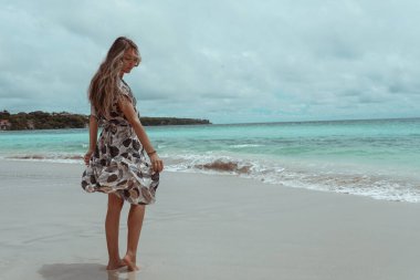 Okyanus ve denizin kıyısında mavi dalgalarla poz veren güzel ve genç bir kız. Uzun elbise mankeni ve moda pozu giymiş bir kadın. Bali adasında rahatla ve rahatla.