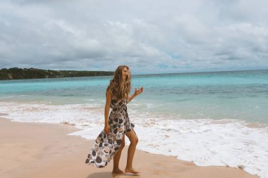 Okyanus ve denizin kıyısında mavi dalgalarla poz veren güzel ve genç bir kız. Uzun elbise mankeni ve moda pozu giymiş bir kadın. Bali adasında rahatla ve rahatla.