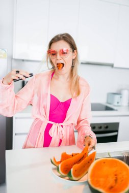 Güzel bir kız mutfakta karpuz keser ve yer. Taze ve sulu karpuz. Pembe mayo ve pelerinli bir kadın. Yaz, tropik bölgeler, Tayland