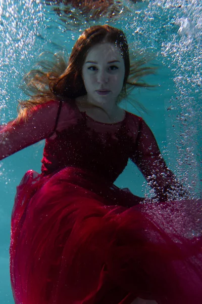 Beautiful girl underwater in a red dress swims in the pool. Tenderness and elegance. Bubbles and a lot of water