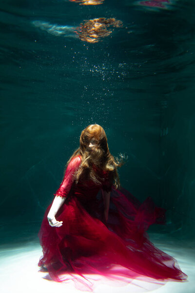 Красивая девушка под водой в красном платье плавает в бассейне. Нежность и элегантность. Пузырьки и много воды
