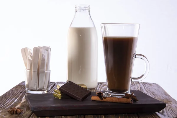 Un vaso de café con chocolate en una bandeja, una botella de leche y bolsas con azúcar en el fondo sobre un fondo blanco . — Foto de Stock