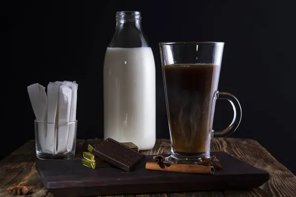 Un vaso de café con chocolate en una bandeja, una botella de leche y bolsas de azúcar en el fondo sobre un fondo negro . — Foto de Stock