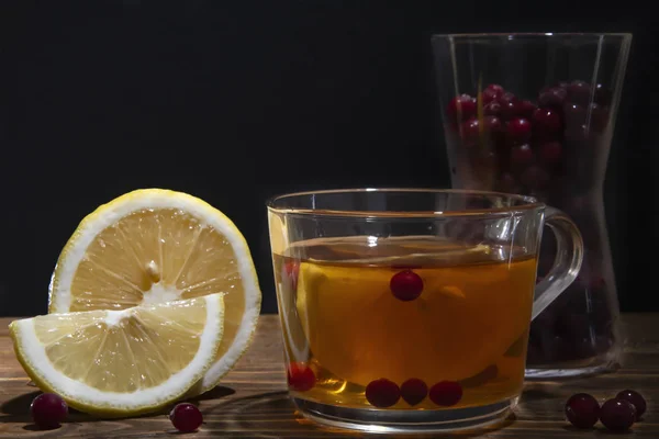 Skleněný šálek s čajem, citronem a brusinkami na dřevěném stole — Stock fotografie